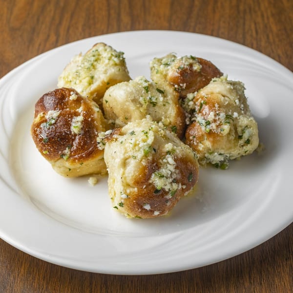 Six golden-brown garlic knots with herbs and cheese on a white plate.